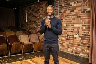 Humoriste noir homme discret sur scène dans un petit club de comédie, tenant un micro avec un sourire subtil et une posture confiante